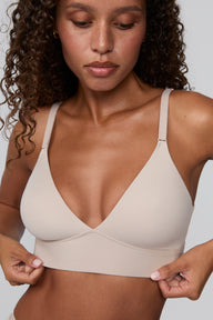 Against a light grey studio backdrop, Courtney wears the Bliss Triangle Bralette in Pebble, looking down while gently holding the bottom band. The bralette features a deep V neckline, wireless lift, and front-adjusting straps in soft microfiber fabric.