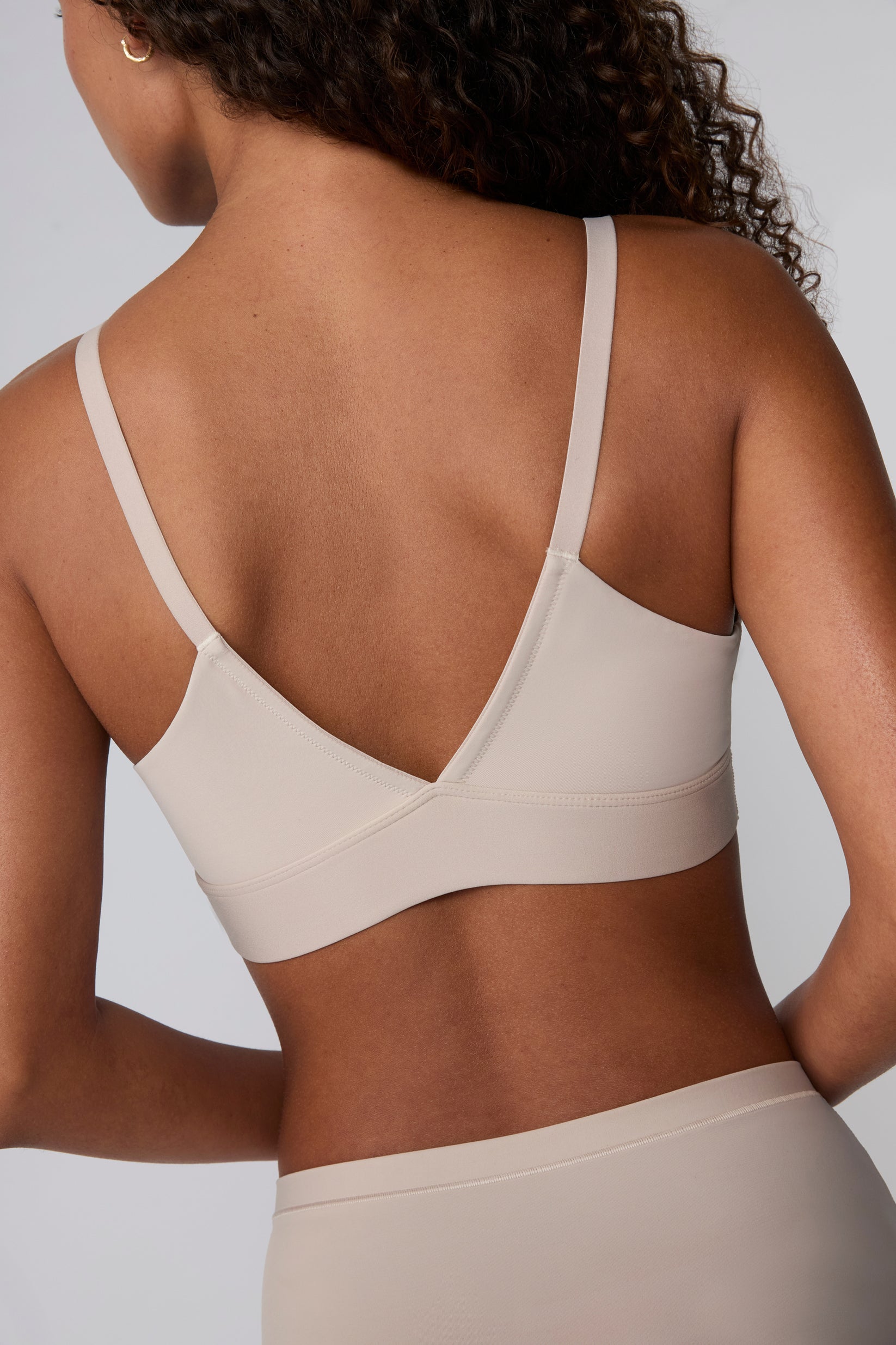 Back view against a light grey studio backdrop, Courtney wears the Bliss Triangle Bralette in Pebble, showing the adjustable straps and wide, smoothing wireless band in soft microfiber fabric.