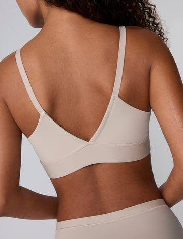 Back view against a light grey studio backdrop, Courtney wears the Bliss Triangle Bralette in Pebble, showing the adjustable straps and wide, smoothing wireless band in soft microfiber fabric.