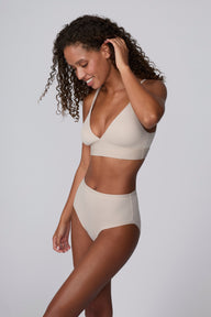 Against a light grey studio backdrop, Courtney wears the Bliss Triangle Bralette in Pebble with matching underwear, looking down with one hand at her hair. The bralette features a deep V neckline, front-adjusting straps, and a wide wireless band in soft microfiber fabric.