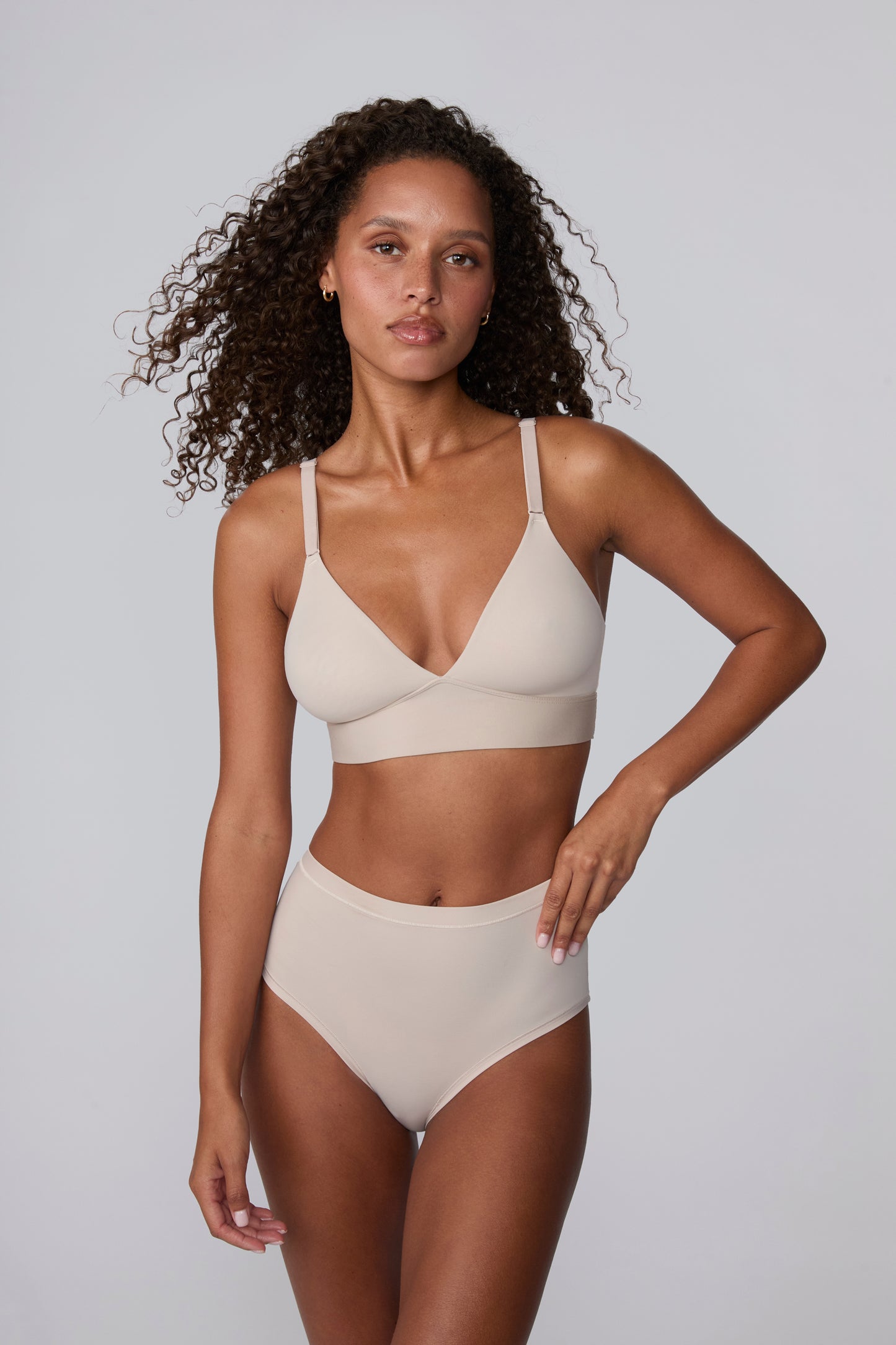 In a light grey studio setting, Courtney wears the Bliss Triangle Bralette in Pebble with matching underwear, standing in a relaxed pose. The deep V front, adjustable straps, and smooth wireless band are visible in soft, breathable microfiber. sizing: Courtney wears size Small