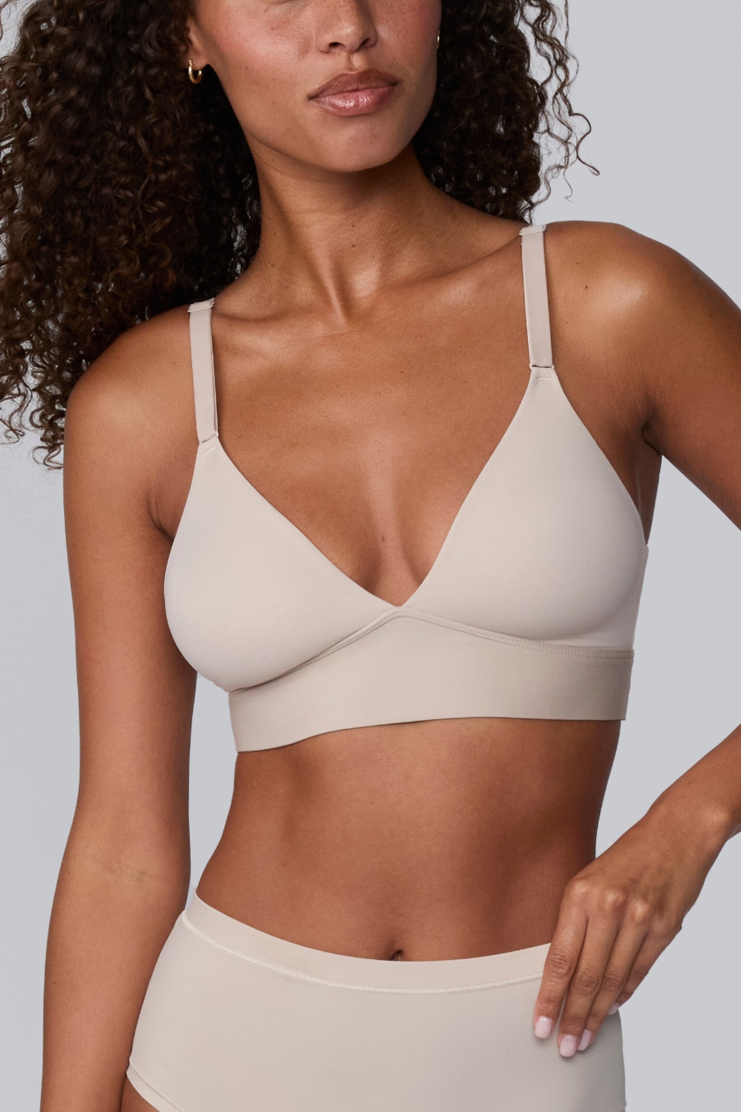 In a light grey studio setting, Courtney wears the Bliss Triangle Bralette in Pebble with matching underwear, standing in a relaxed pose. The deep V front, adjustable straps, and smooth wireless band are visible in soft, breathable microfiber. sizing: Courtney wears size Small