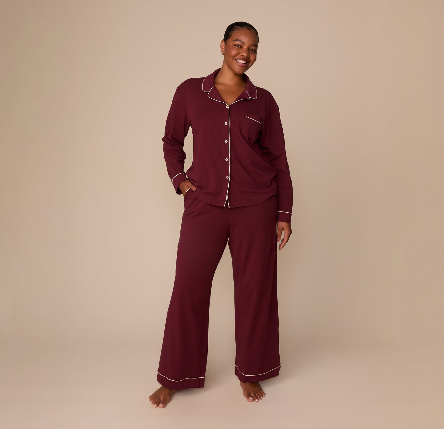 Full-body view of model Zarria wearing the Perfect Pima Long Sleeve Pajama Shirt and Pants in Bordeaux. She stands barefoot with one hand in her pocket, smiling softly.