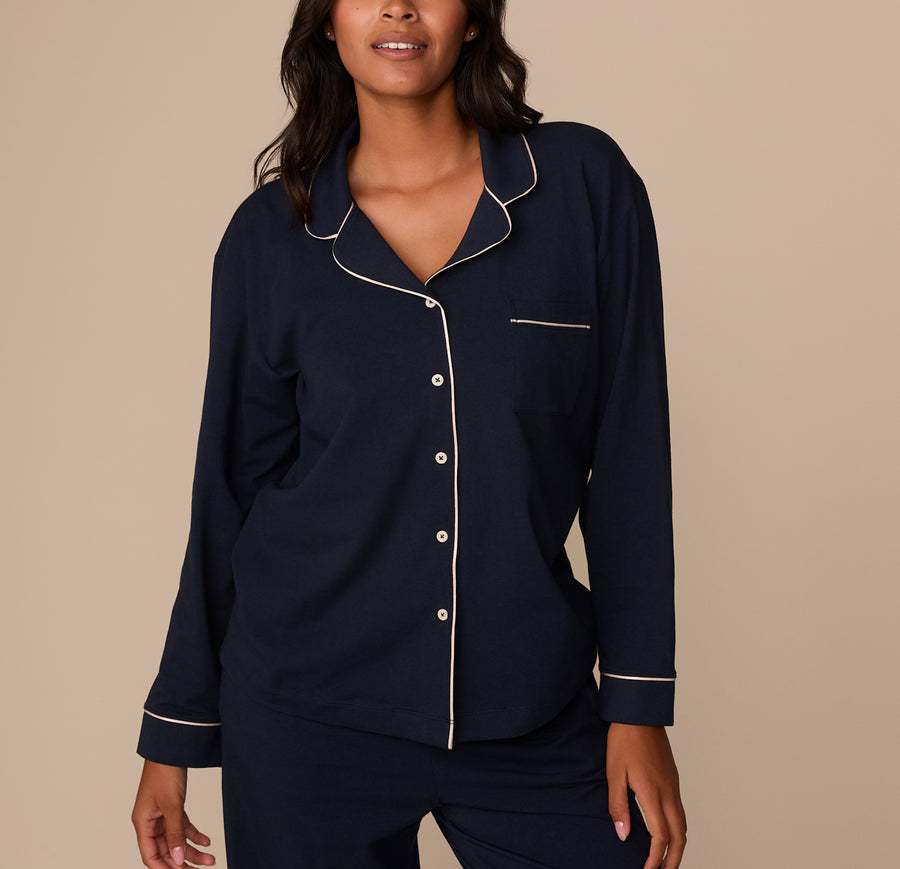 Victoria wears the Perfect Pima Long Sleeve Pajama Shirt in deep navy blue. The shirt features a classic notched collar, white contrast piping along the collar, front placket, pocket, and cuffs, and is styled with white buttons. She stands confidently with a slight smile, one hand relaxed at her side and the other partially tucked into her matching pajama pants. sizing: Victoria wears size Medium