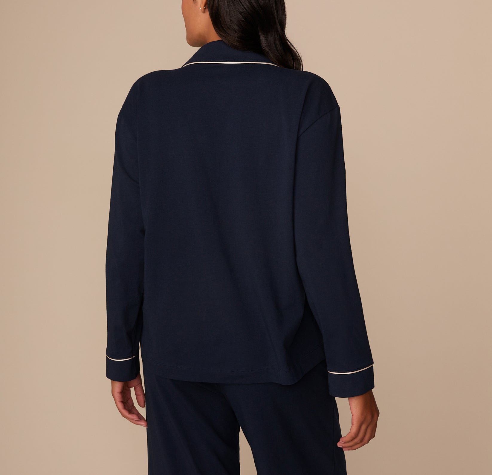 Back view of Victoria modeling the pajama shirt, showcasing the clean, seamless back panel of the shirt. The white piping along the collar and sleeve cuffs stands out against the solid navy fabric. Her dark hair drapes down naturally over the back, complementing the soft, relaxed silhouette of the shirt.