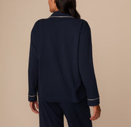 Back view of Victoria modeling the pajama shirt, showcasing the clean, seamless back panel of the shirt. The white piping along the collar and sleeve cuffs stands out against the solid navy fabric. Her dark hair drapes down naturally over the back, complementing the soft, relaxed silhouette of the shirt.