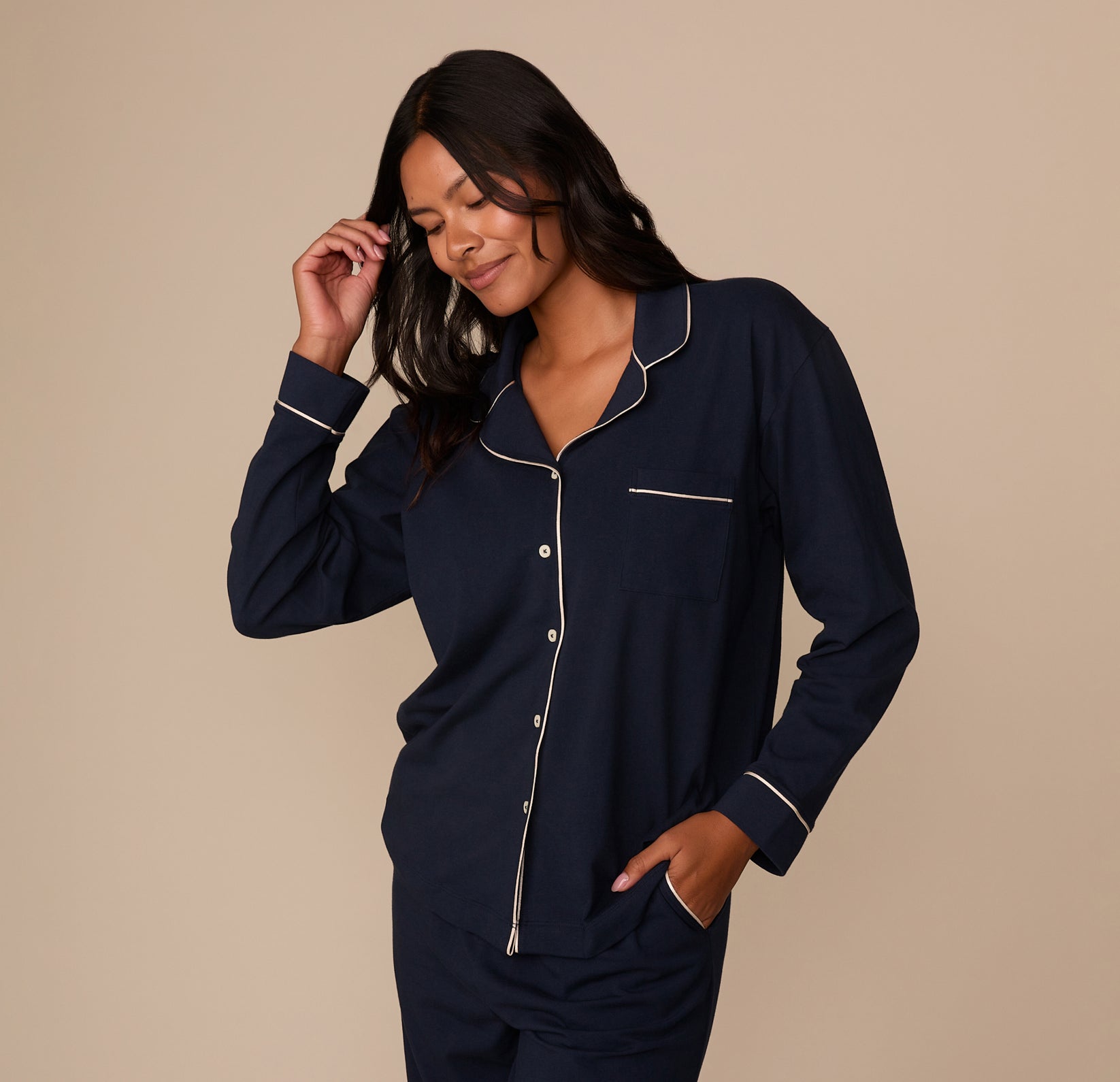 Victoria is shown in a three-quarter profile, gently smiling with one hand touching her temple and the other in her pocket. The shirt's relaxed drape, long sleeves, and classic pajama styling are visible. The curved hem and tailored yet loose fit emphasize comfort and sophistication.