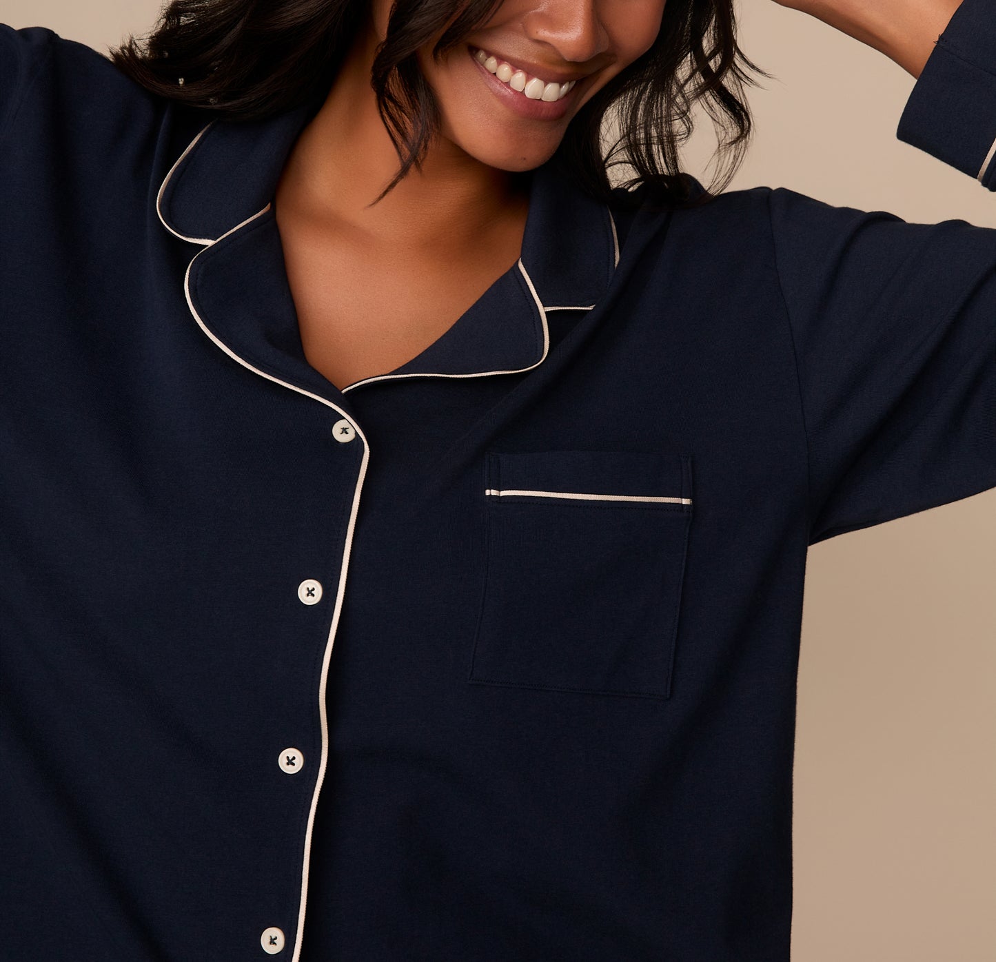 A close-up of the upper half of the pajama shirt focuses on the collar, front buttons, and chest pocket. The white piping trims the edge of the pocket and collar with precision, and the soft, fine-knit texture of the Pima cotton is evident. Victoria’s smile and relaxed posture enhance the cozy, elegant vibe.