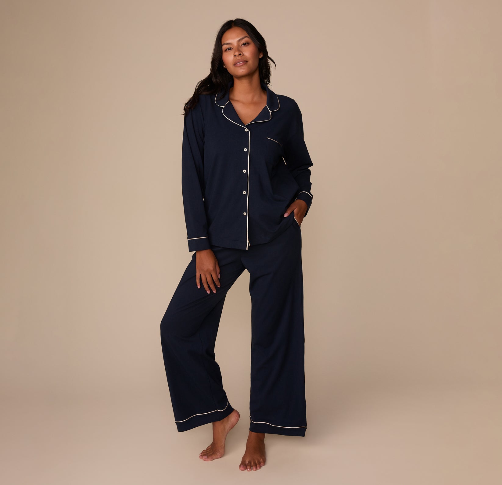 Victoria models the full pajama set, including the matching Navy Pants with white piping at the cuffs. She stands barefoot on a neutral beige background, slightly leaning with a relaxed, casual stance. One hand rests in her pant pocket while the other hangs by her side, highlighting the drape and fit of both the shirt and pants as a coordinated set.