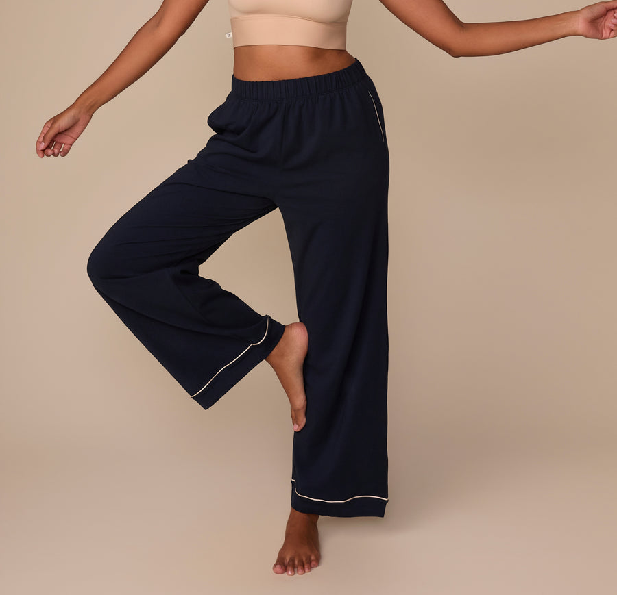 Victoria playfully balances on one leg while wearing the Perfect Pima Pajama Pants in Navy. The pants feature a relaxed wide-leg silhouette with contrast ivory piping at the cuffs, and an elastic waistband for comfort. sizing: Victoria wears size Medium