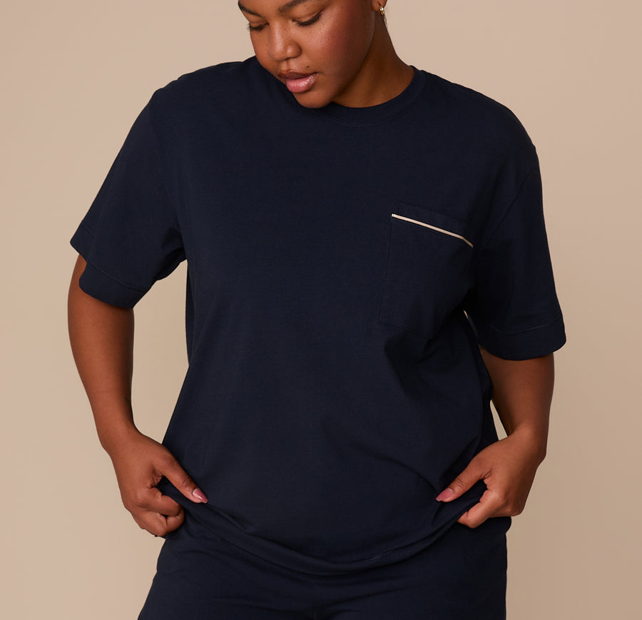 Zarria wears the Perfect Pima Pajama Tee in navy, standing with hands near her hips. The relaxed-fit tee features a crew neckline, short sleeves, and a chest pocket with ivory contrast piping detail. She pairs it with matching pajama shorts. sizing: Zarria wears size XL