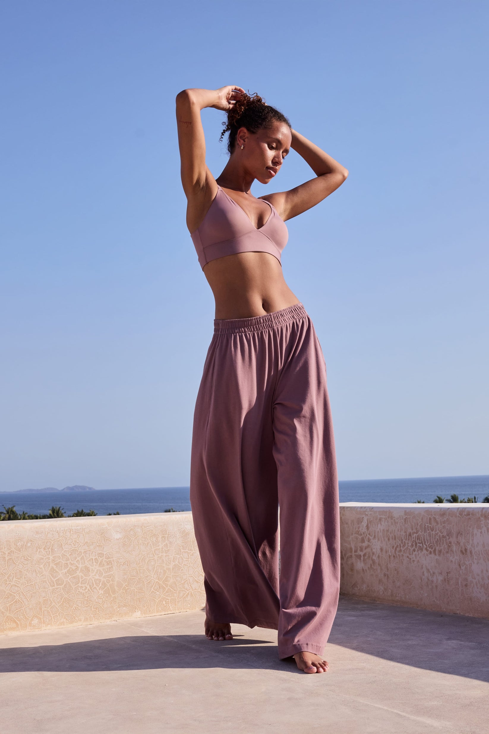 On a sunlit terrace overlooking the ocean, Courtney wears the Pima Palazzo Pant in Mirage with a matching wireless bra, showing the relaxed wide-leg silhouette, elastic waistband, and soft drape.