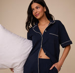 Marcela wears the Perfect Pima Pajama Shirt in Navy, styled playfully with a white pillow tucked under one arm and the shirt unbuttoned slightly at the waist to show the matching shorts' elastic waistband.