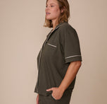 Side profile of Sonnie in the Perfect Pima Pajama Shirt in Olive, with one hand in the pocket of the matching Perfect Pima Pajama Shorts and white piping visible on the sleeve and chest pocket.