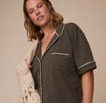 Sonnie wears the Perfect Pima Pajama Shirt in Olive, styled with a cozy cream knit throw draped over her shoulder, highlighting the shirt’s soft fabric and contrast piping.