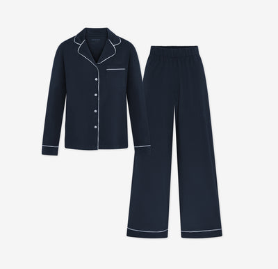 Harper Wilde Pima Pajama Set in Navy. Includes long sleeve, button-up top with white piping and matching long pajama pants. Set laid flat on a white background.