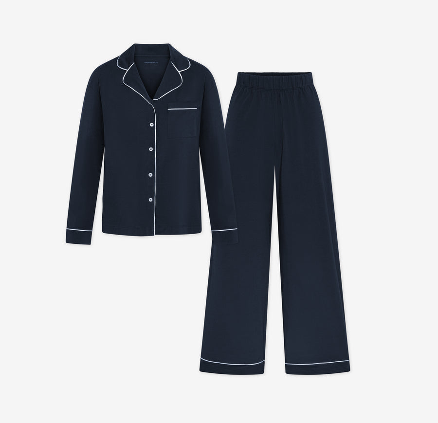 Harper Wilde Pima Pajama Set in Navy. Includes long sleeve, button-up top with white piping and matching long pajama pants. Set laid flat on a white background.