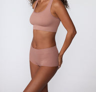 On a white studio backdrop, a model wears the Ribbed Boyshort in Plum, shown from a side angle. The soft ribbed fabric and smooth waistband sit comfortably at the hips with full coverage through the back.