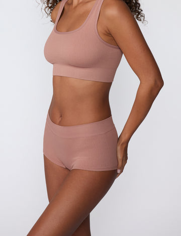 On a white studio backdrop, a model wears the Ribbed Boyshort in Plum, shown from a side angle. The soft ribbed fabric and smooth waistband sit comfortably at the hips with full coverage through the back.