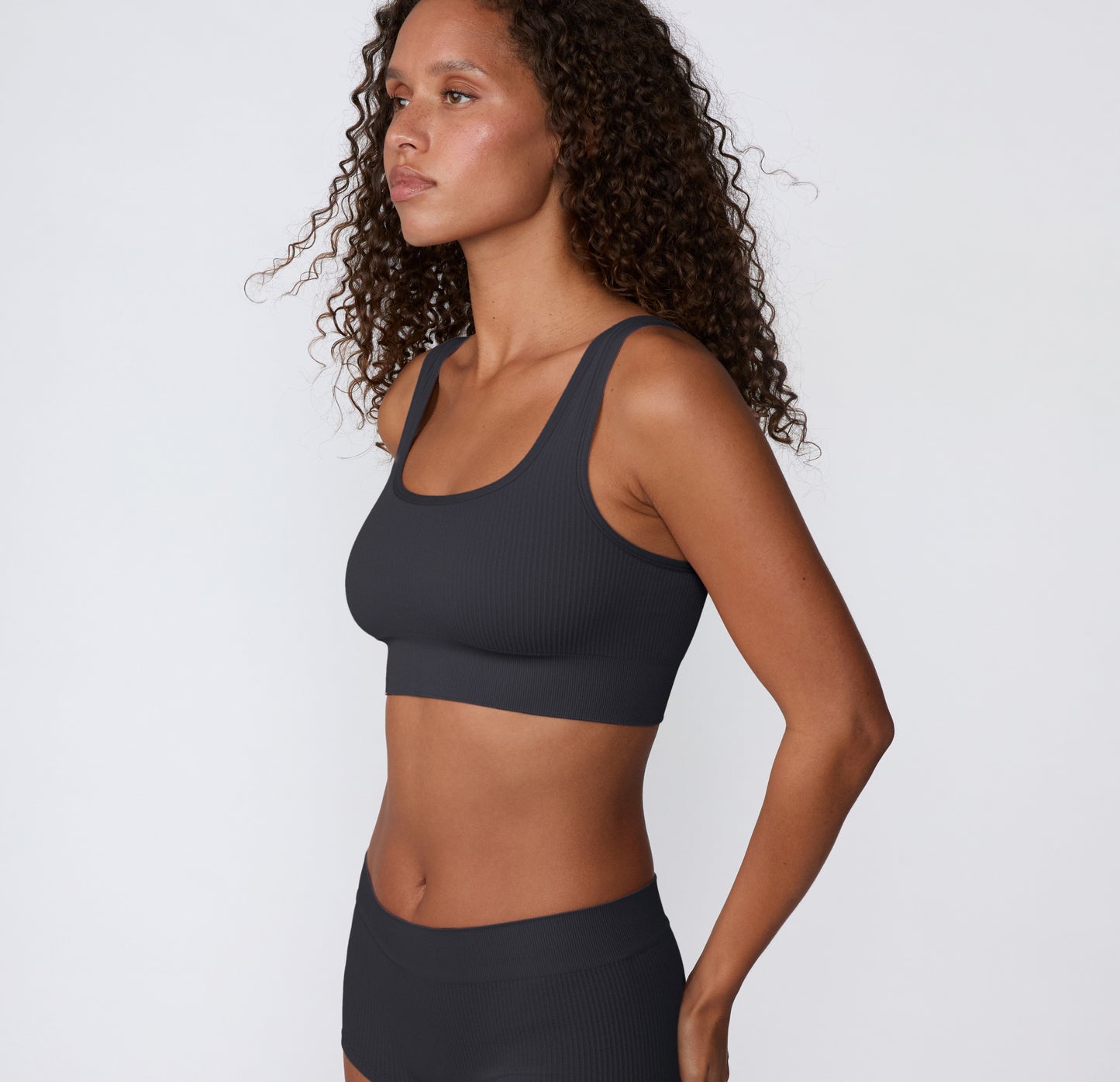 On a light studio backdrop, a side profile shows a model wearing the Ribbed Scoop Bralette in Charcoal, featuring the scoop neckline, smooth ribbed texture, and supportive underband.