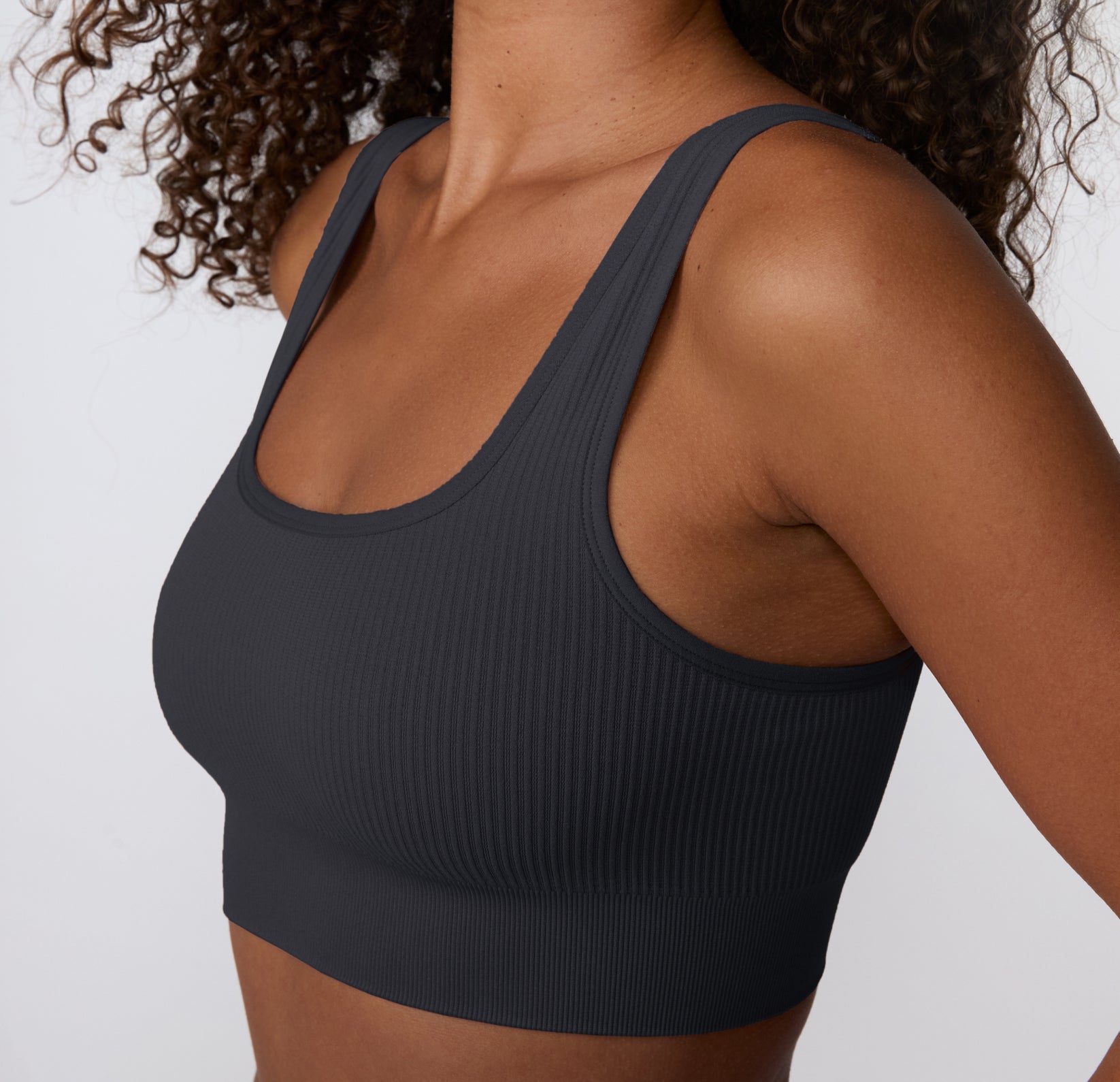 On a light studio backdrop, a close-up angled view shows a model wearing the Ribbed Scoop Bralette in Charcoal, focusing on the scoop neckline, ribbed texture, and soft, stretchy fabric.