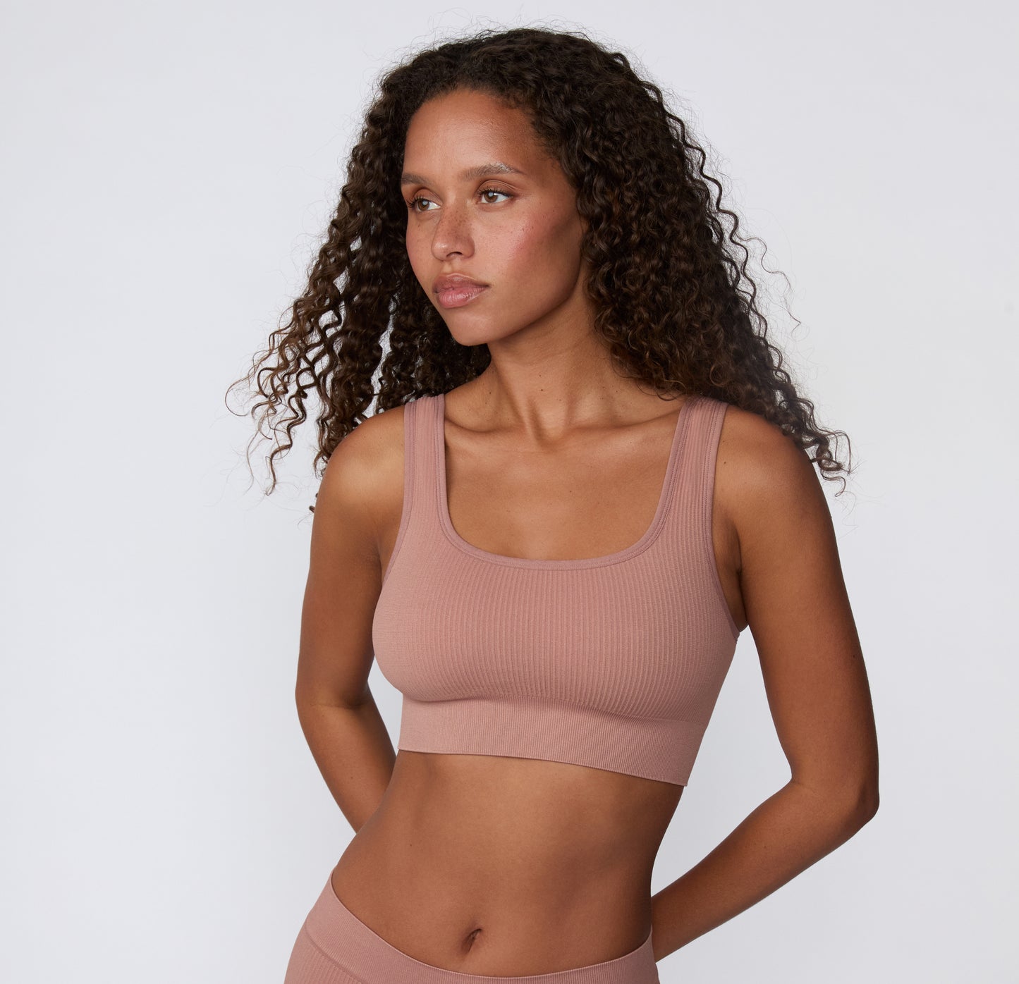 On a light studio backdrop, a three-quarter front view shows a model wearing the Ribbed Scoop Bralette in Plum, highlighting the scoop neckline, soft ribbed fabric, and wireless construction.