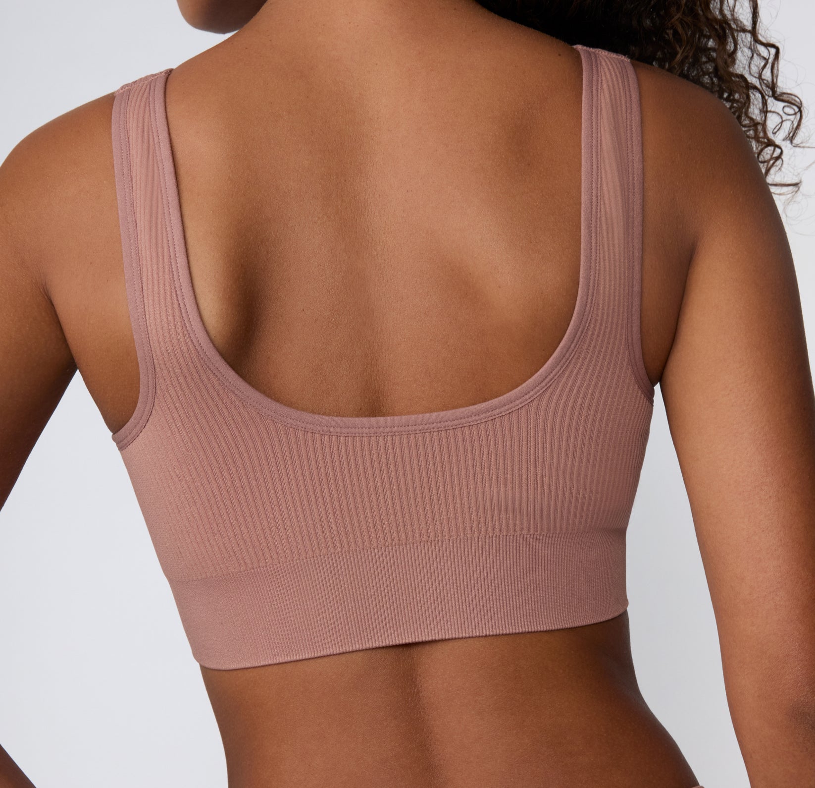 On a light studio backdrop, a back view shows a model wearing the Ribbed Scoop Bralette in Plum, highlighting the scoop back shape, wide straps, and smooth ribbed fabric.