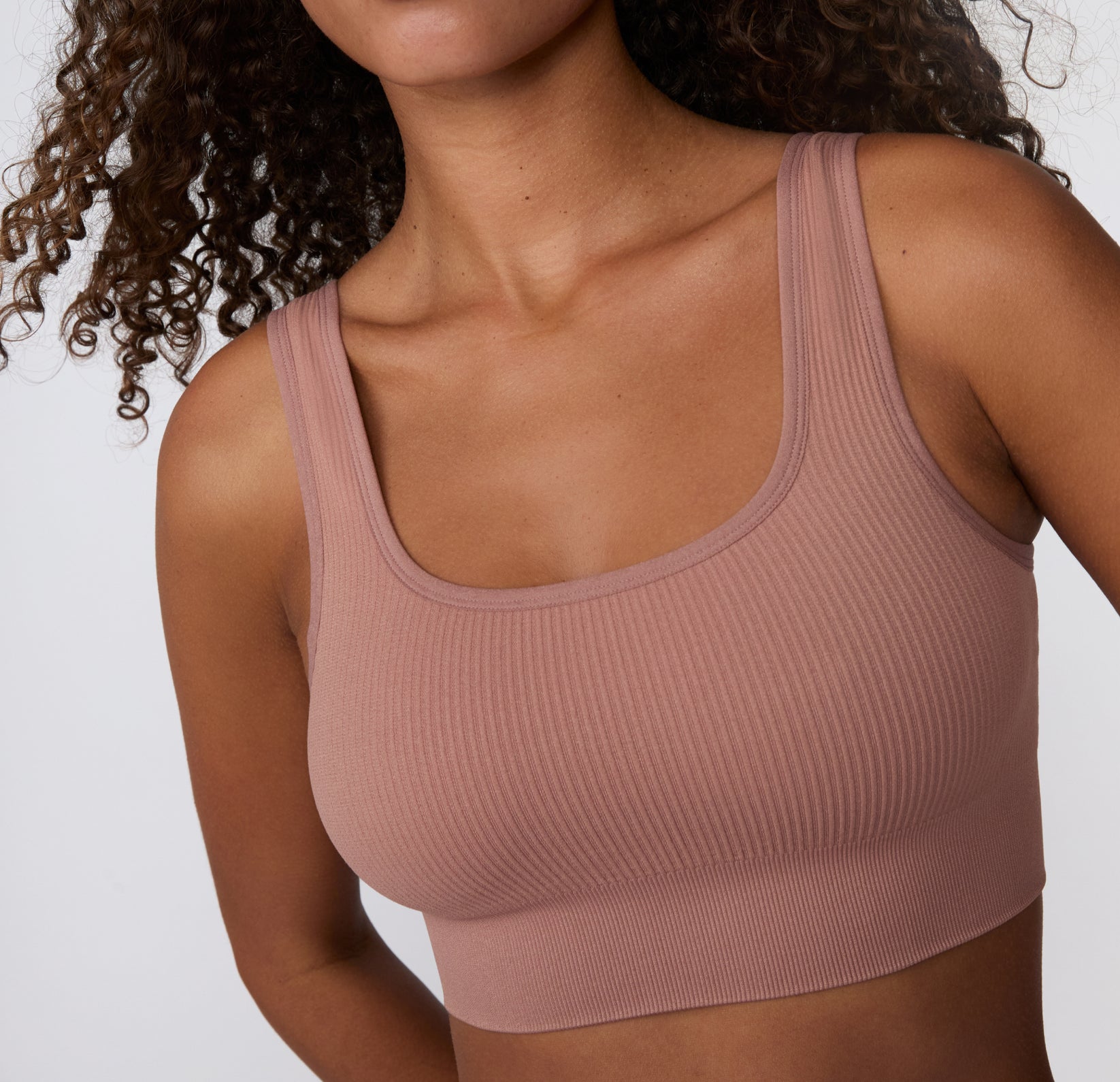 On a light studio backdrop, a close-up angled view shows a model wearing the Ribbed Scoop Bralette in Plum, focusing on the scoop neckline, ribbed texture, and soft, stretchy fabric.