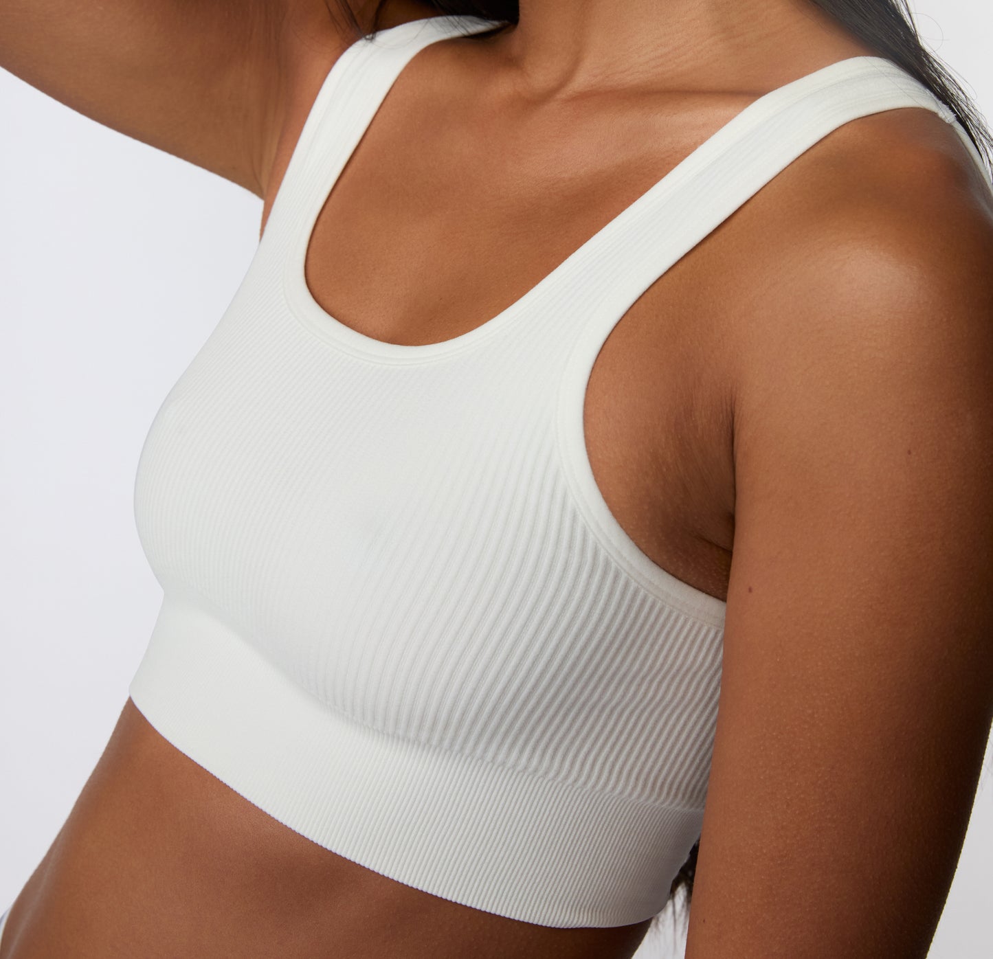 On a light studio backdrop, a close-up angled view shows a model wearing the Ribbed Scoop Bralette in White, focusing on the ribbed texture, smooth underband, and wireless design.