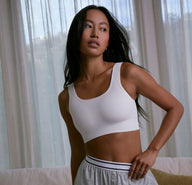 In a softly lit room with sheer curtains, a model wears the Ribbed Scoop Bralette in White, styled with lounge shorts to show the comfortable, everyday fit.