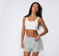 On a light studio backdrop, a full-body front view shows a model wearing the Ribbed Scoop Bralette in White with lounge shorts, emphasizing the scoop neckline and ribbed fabric.