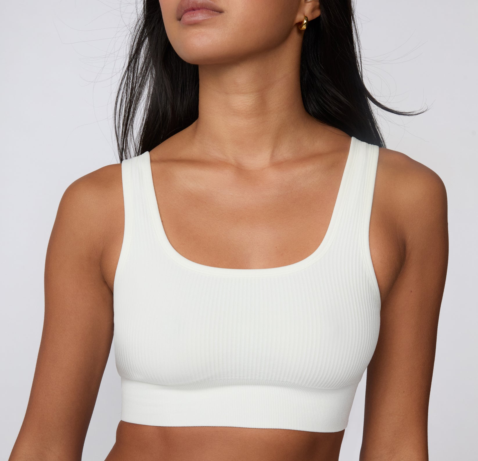 On a light studio backdrop, a close-up front view shows a model wearing the Ribbed Scoop Bralette in White, highlighting the scoop neckline, ribbed texture, and soft stretch.