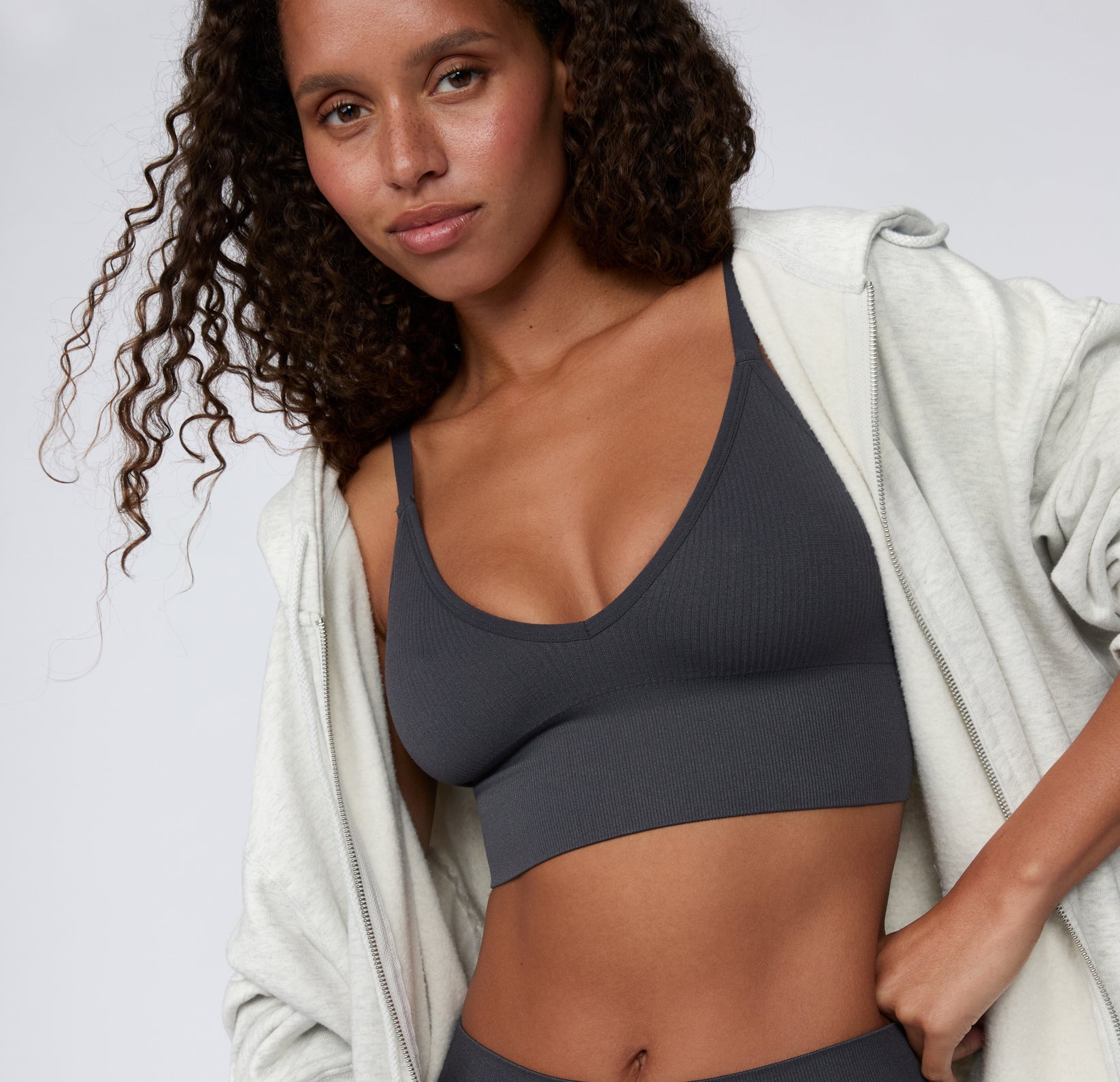 On a light studio backdrop, a close-up front view shows a model wearing the Ribbed Triangle Bralette in Charcoal, highlighting the triangle neckline, soft ribbed fabric, and thin straps, styled under an open zip hoodie.