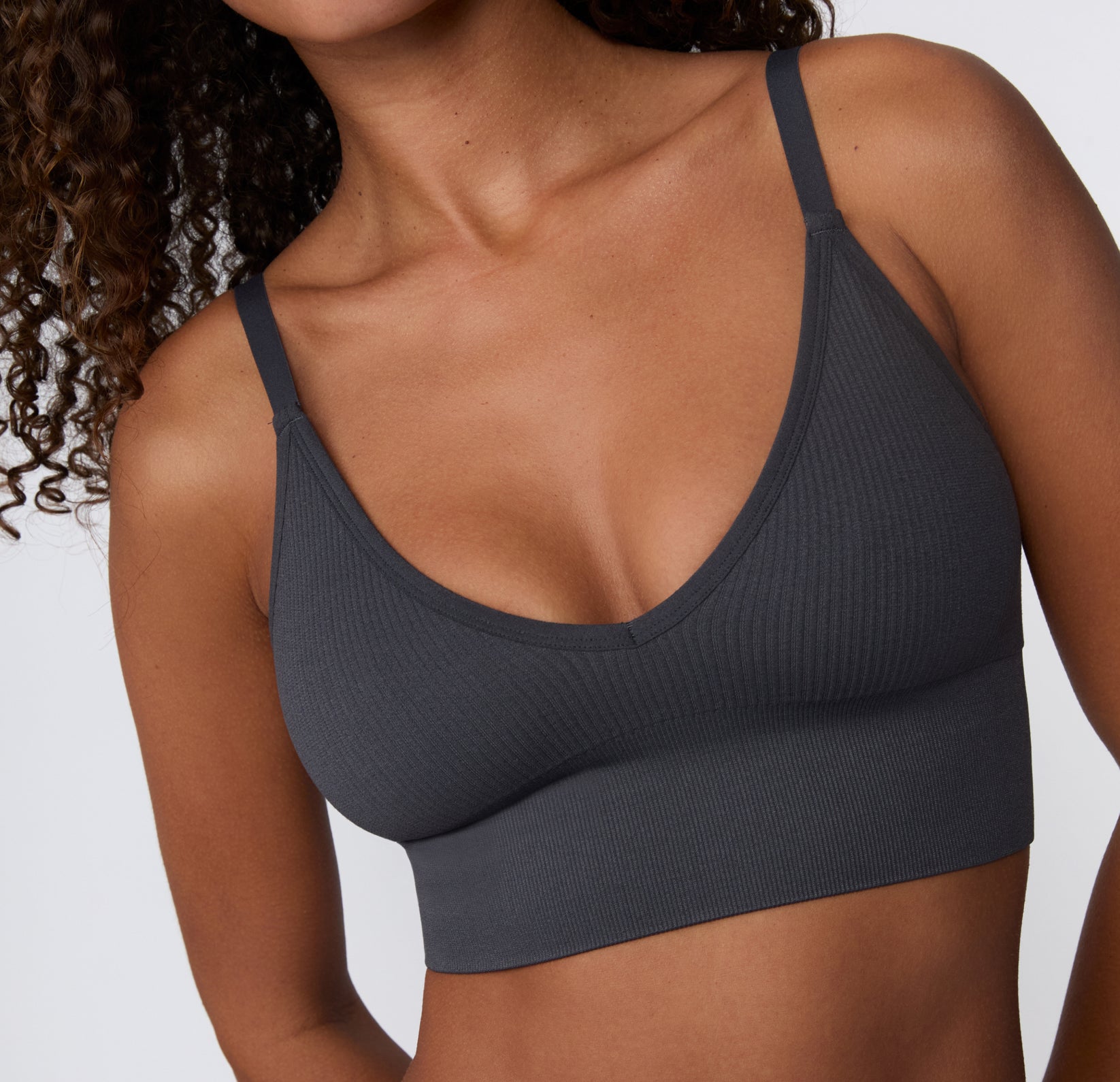 On a light studio backdrop, a close-up angled view shows a model wearing the Ribbed Triangle Bralette in Charcoal, focusing on the triangle neckline, ribbed fabric, and smooth underband.