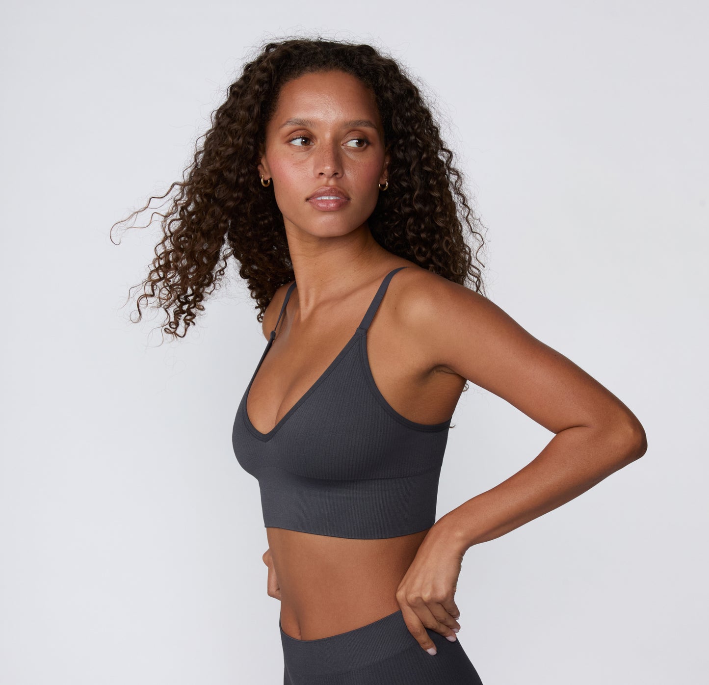 On a light studio backdrop, a three-quarter side view shows a model wearing the Ribbed Triangle Bralette in Charcoal, highlighting the slim straps and ribbed construction.