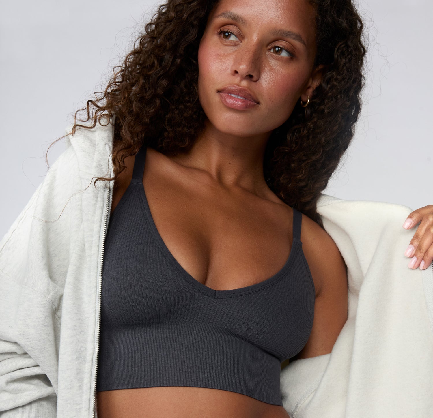 On a light studio backdrop, a styled close-up shows a model wearing the Ribbed Triangle Bralette in Charcoal layered under a zip hoodie, showing the triangle neckline and ribbed fabric. sizing: Courtney wears size Small