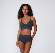 On a light studio backdrop, a full-body front view shows a model wearing the Ribbed Triangle Bralette with matching underwear in Charcoal, emphasizing the ribbed fabric and wireless design.