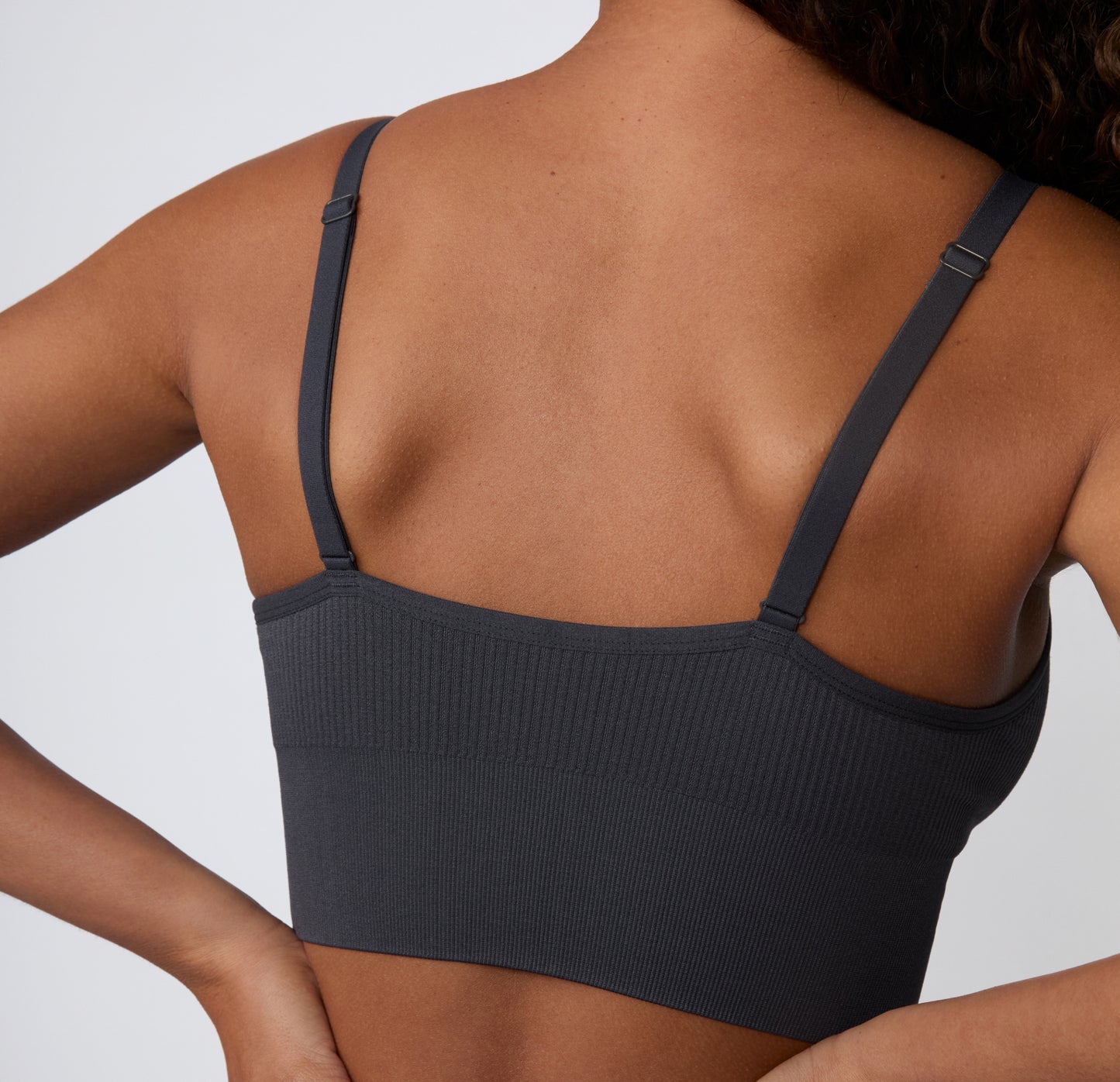 On a light studio backdrop, a close-up angled view shows a model wearing the Ribbed Triangle Bralette in Charcoal, focusing on the ribbed texture, triangle shape, and soft stretch.