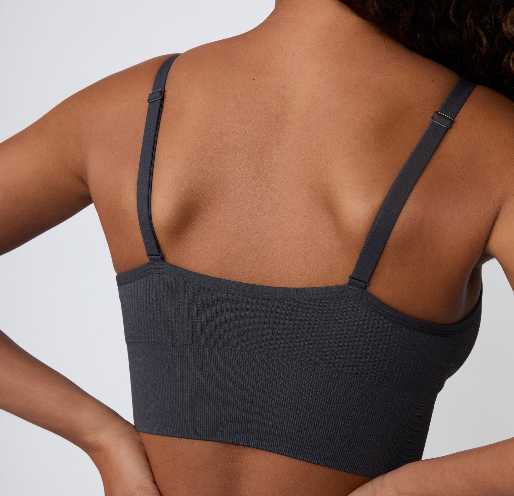On a light studio backdrop, a close-up angled view shows a model wearing the Ribbed Triangle Bralette in Charcoal, focusing on the ribbed texture, triangle shape, and soft stretch.