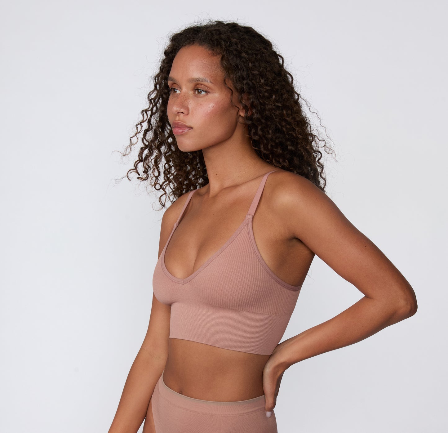 On a light studio backdrop, a three-quarter side view shows a model wearing the Ribbed Triangle Bralette in Plum, highlighting the slim straps, ribbed construction, and soft stretch.