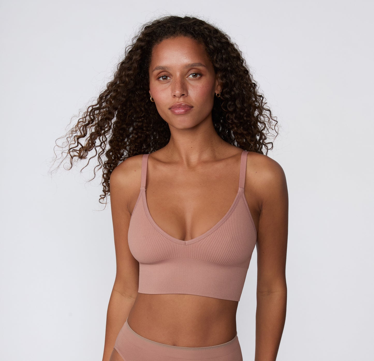 On a light studio backdrop, a front-facing mid-length view shows a model wearing the Ribbed Triangle Bralette in Plum, highlighting the triangle neckline, ribbed fabric, and smooth underband.