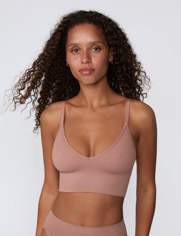 On a light studio backdrop, a front-facing mid-length view shows a model wearing the Ribbed Triangle Bralette in Plum, highlighting the triangle neckline, ribbed fabric, and smooth underband.
