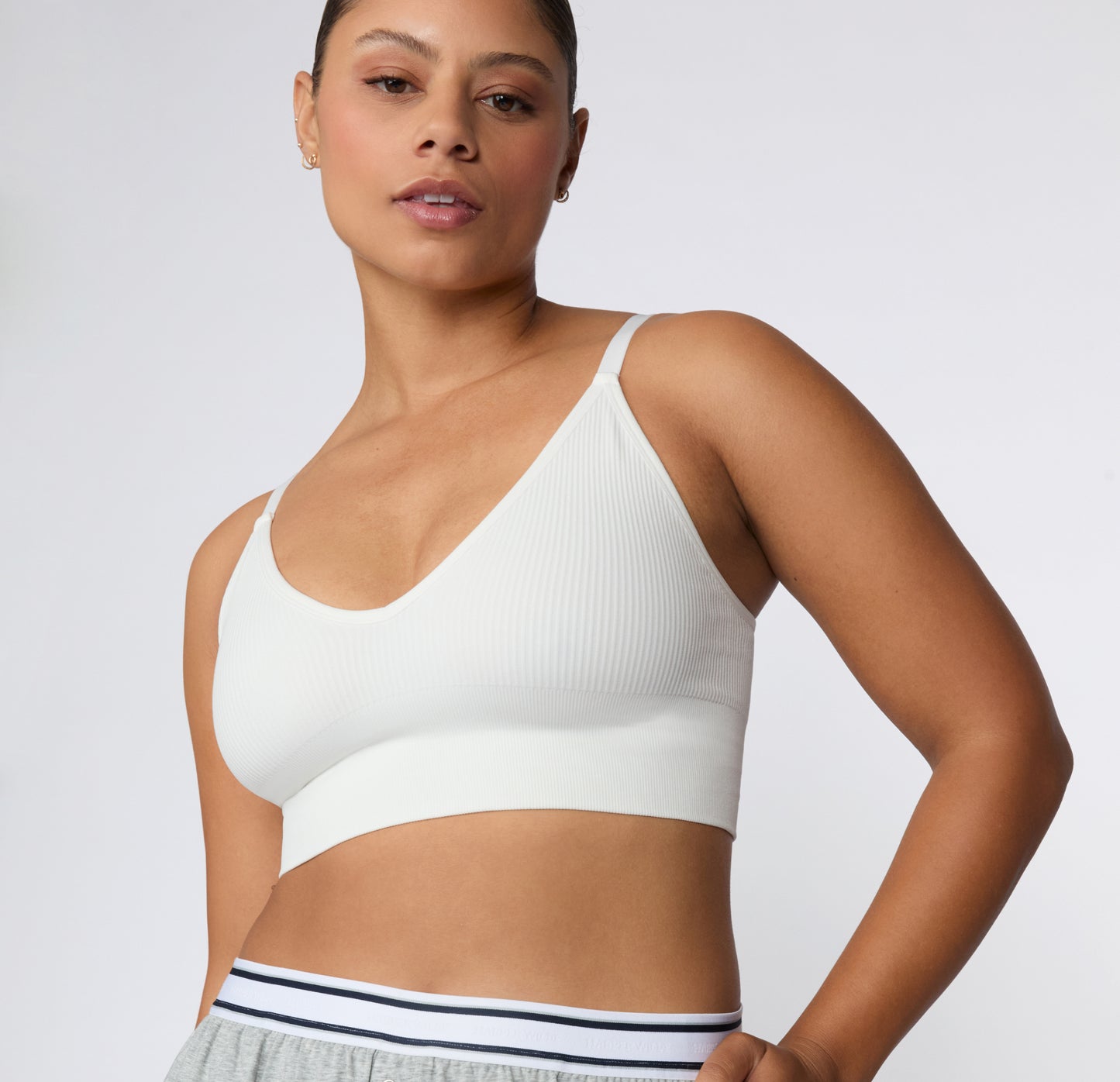 On a light studio backdrop, a close-up angled front view shows a model wearing the Ribbed Triangle Bralette in White, highlighting the triangle neckline, ribbed fabric, and thin straps.