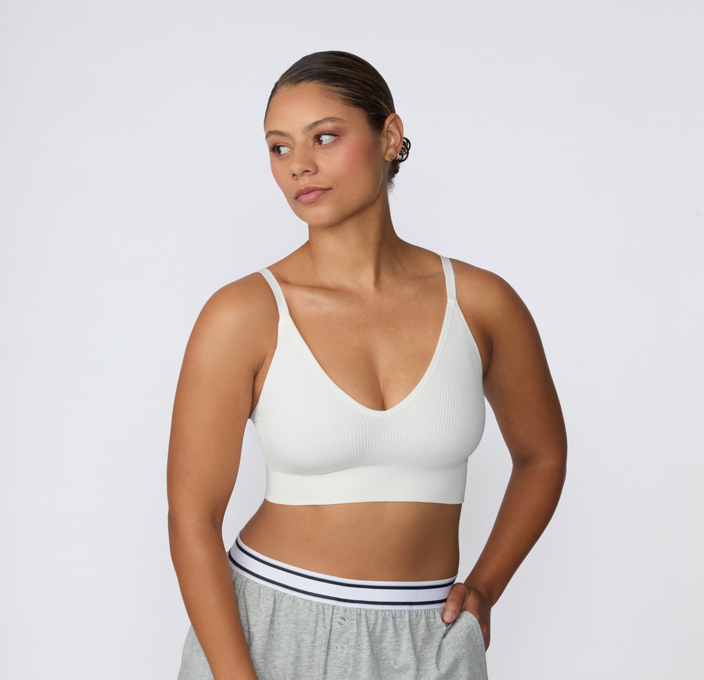 On a light studio backdrop, a three-quarter front view shows a model wearing the Ribbed Triangle Bralette in White with lounge shorts, highlighting the triangle neckline, ribbed construction, and thin straps.