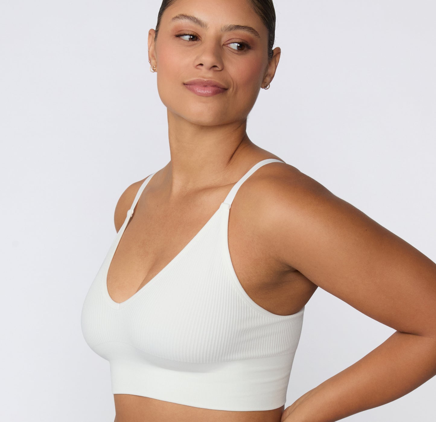 On a light studio backdrop, a three-quarter side view shows a model wearing the Ribbed Triangle Bralette in White, highlighting the triangle neckline, ribbed fabric, and thin straps.