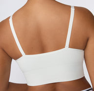 On a light studio backdrop, a close-up back view shows a model wearing the Ribbed Triangle Bralette in White, focusing on the straight back shape, adjustable straps, and ribbed texture.