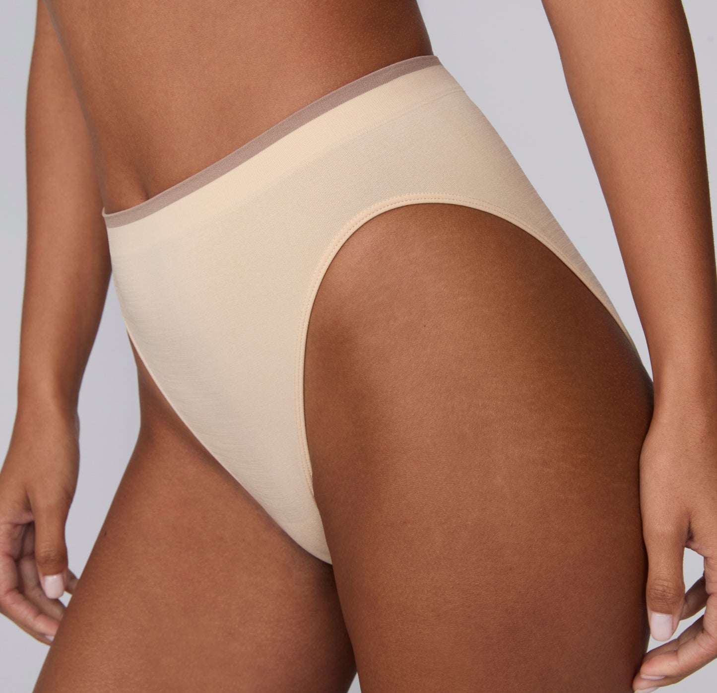 On a light gray studio backdrop, a side close-up of Courtney wearing the Smooth Seamless underwear in Beige shows the smooth, stretchy fabric and flat edges along the leg opening.