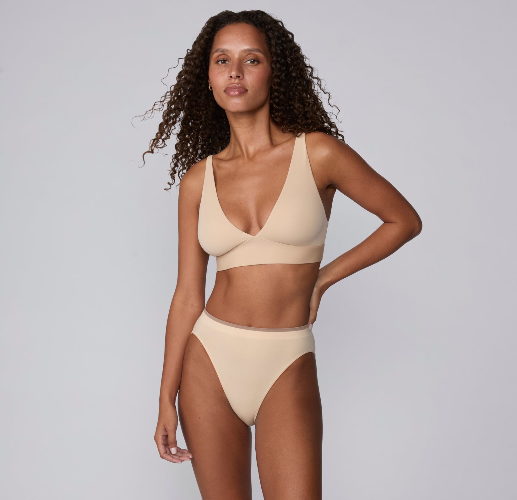 On a light gray studio backdrop, Courtney wears the Smooth Seamless underwear in Beige in a full-body front view, paired with a matching bra to show the low-rise fit and smooth waistband.