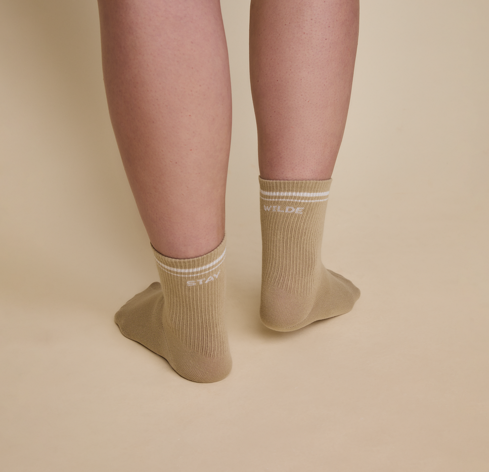 Close up of a model wearing the Quarter Crew Socks in the color Tarragon. The back of the left sock says "Stay" and the back of the right sock says "Wilde".