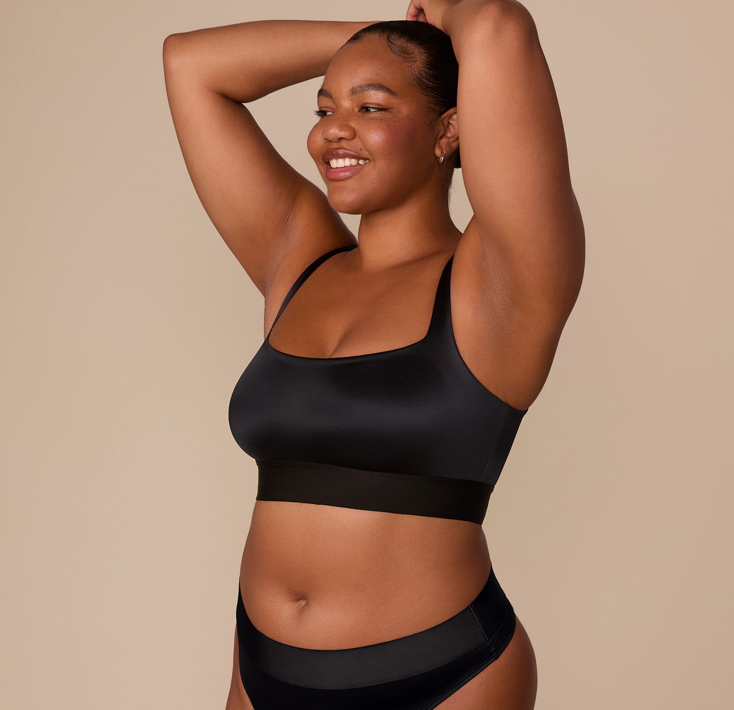 Zarria smiling with arms raised, wearing the Stretch Satin Square Neck Bralette in Black. The satin finish and extended underband are clearly visible.